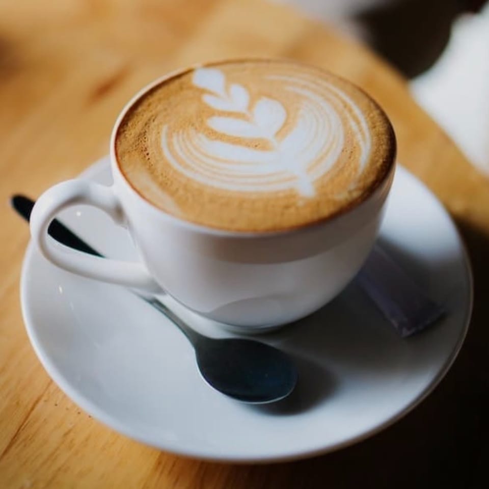 A white cup of latte with a leaf design in the foam, on a saucer with a spoon.